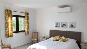Ein Schlafzimmer mit einem Bett, einem Sessel, einem Nachttisch, einer Klimaanlage und einem Fenster mit Vorhängen.