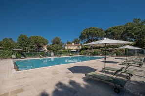 A swimming pool with lounge chairs and umbrellas, surrounded by greenery and a building in the background.