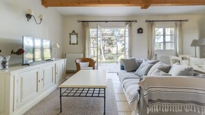 Un salon spacieux avec une grande fenêtre, un canapé blanc, une table basse et un téléviseur posé sur un meuble.