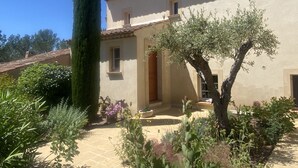 Un bâtiment avec un toit recouvert de tuiles, une porte et une fenêtre. On peut voir des plantes et un grand arbre devant le bâtiment.
