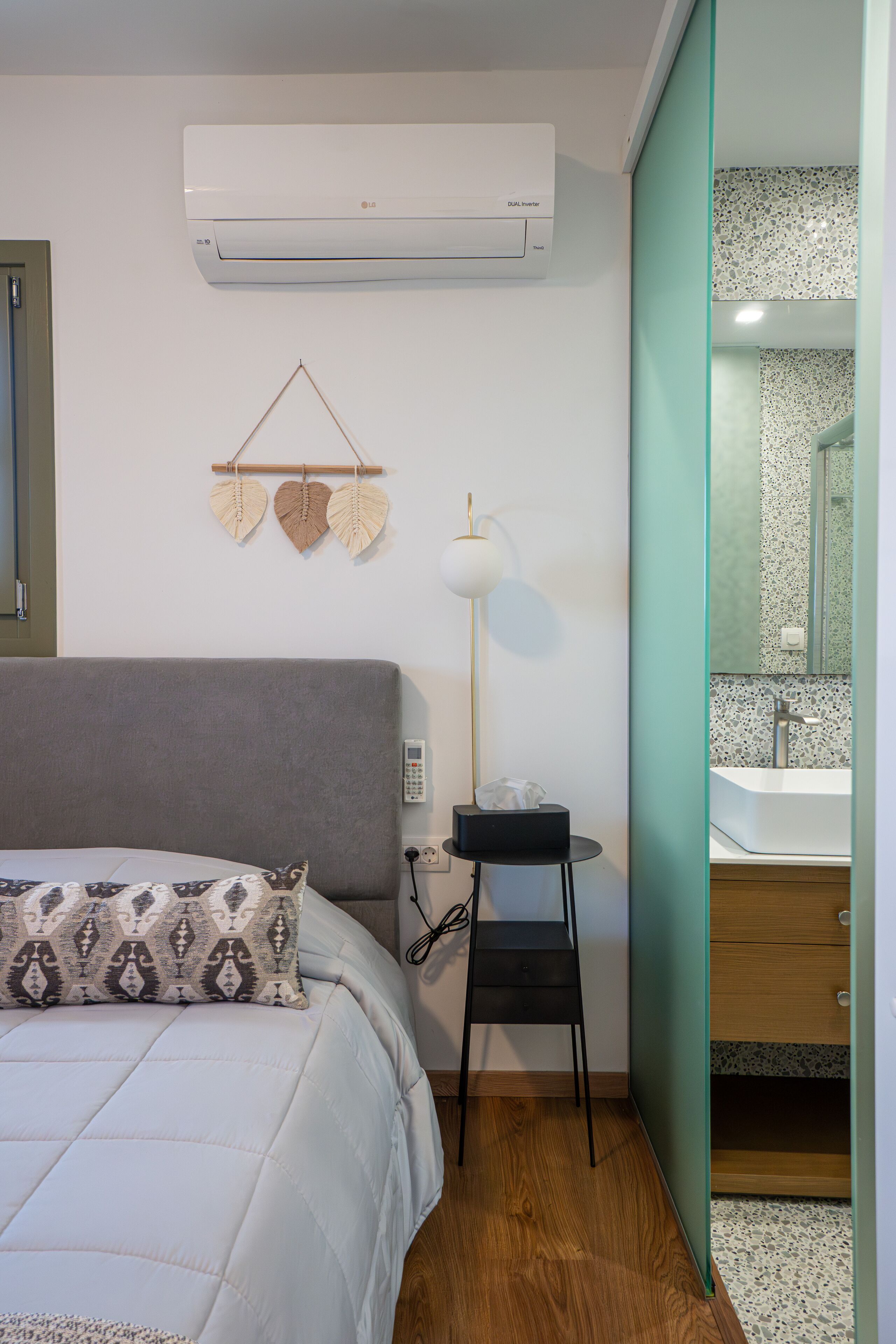 A bedroom with a bed, an air conditioner, a bedside table with a lamp, and a bathroom visible through a glass door.