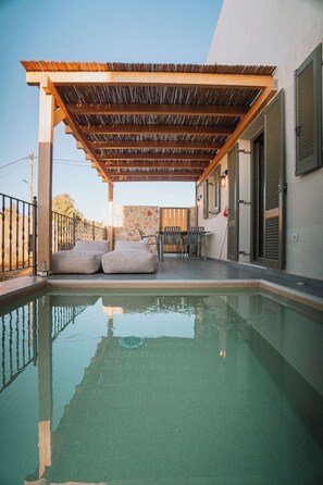 A swimming pool with a wooden pergola overhead, outdoor seating, and a view of a building with shutters.