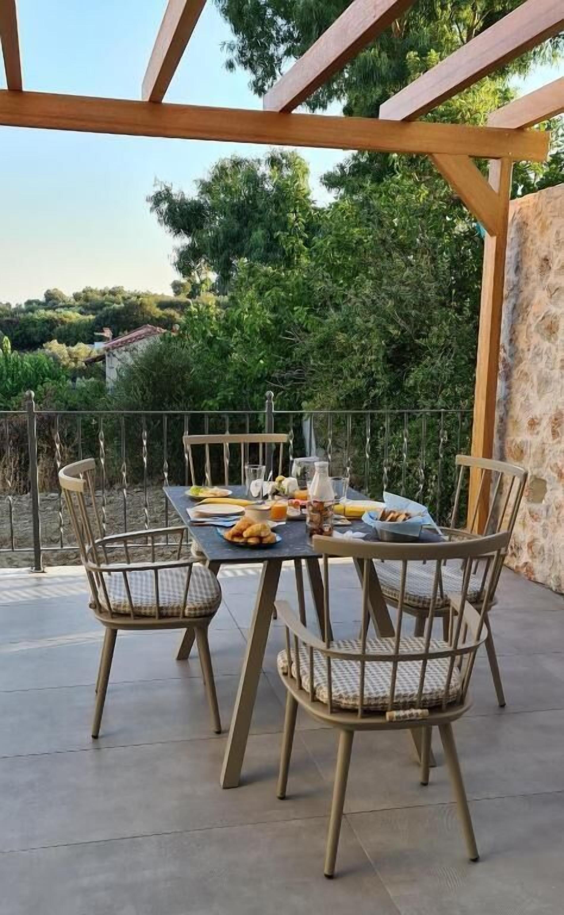 An outdoor dining area with a table set for two, surrounded by wicker chairs, under a wooden pergola.