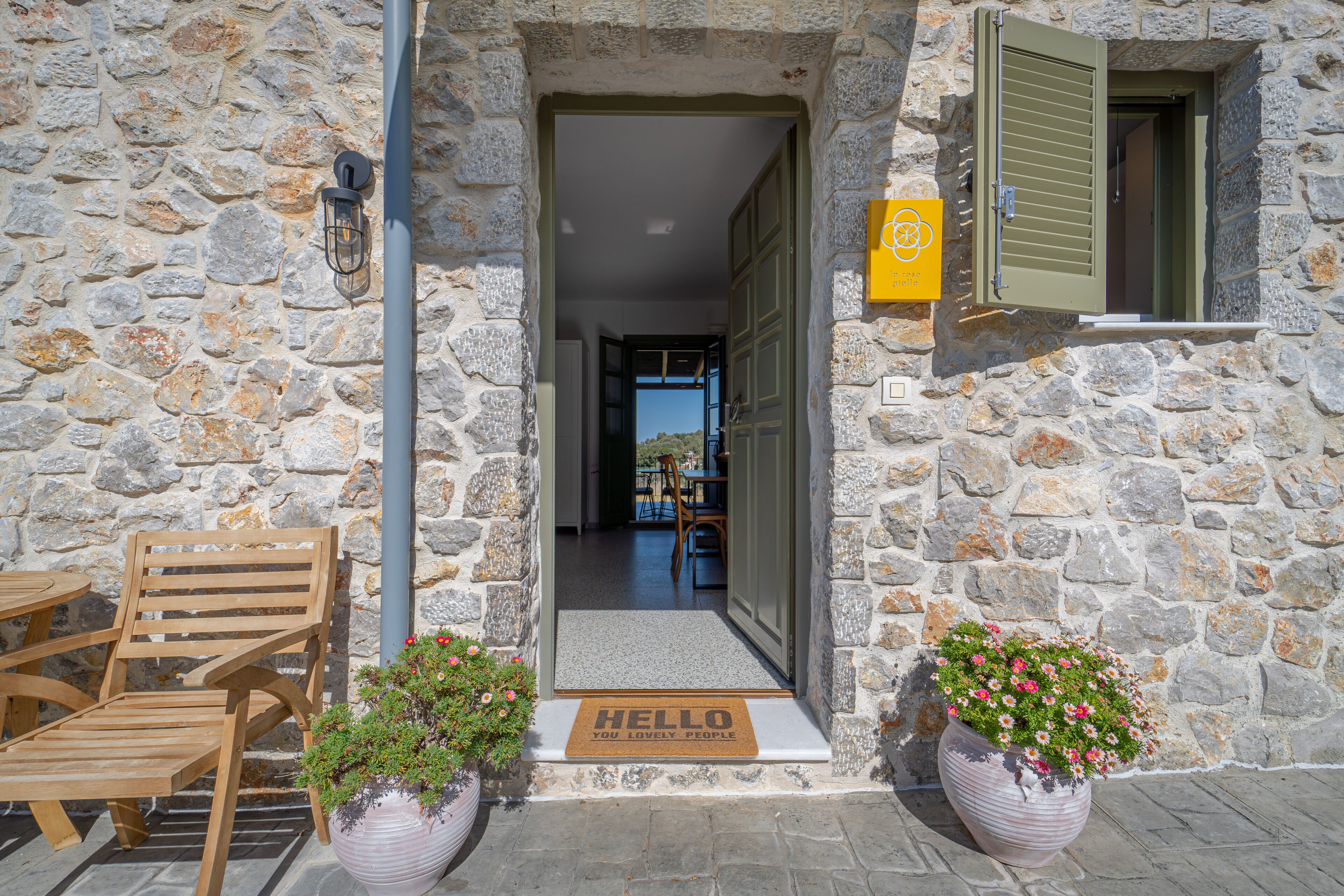 A stone building entrance with a wooden bench, a doormat, and potted plants.
