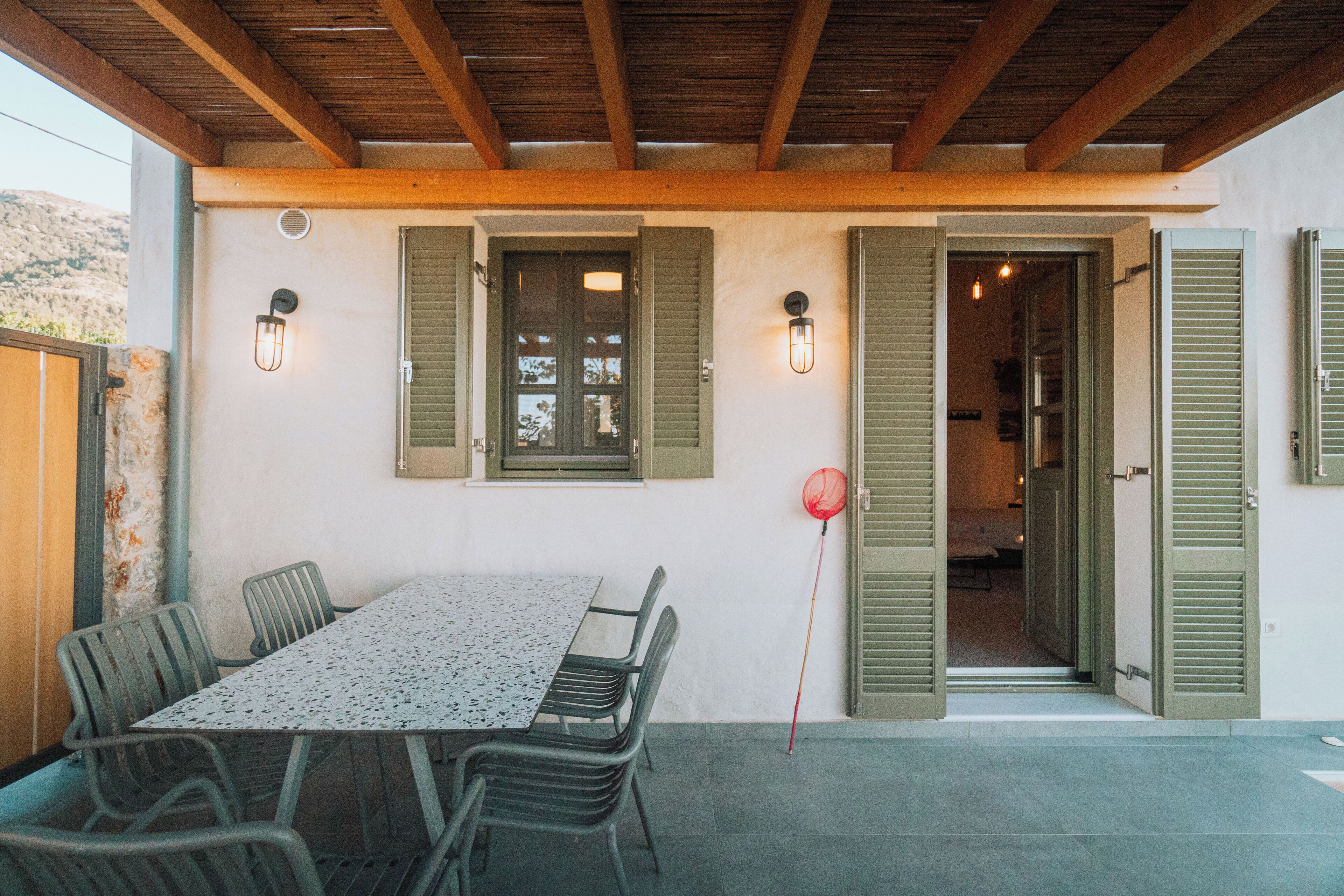 An outdoor patio with a table and chairs, wooden beams overhead, and green shutters on the walls.