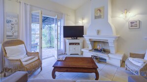 A spacious living room with a television, fireplace, wicker furniture, and a view through the open French doors. - 2 BEDROOM TOWNHOUSE WITH GARDEN (N°507) (La Motte)