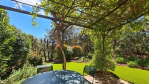 Un patio exterior con una mesa redonda y sillas, rodeado de abundante vegetación y un césped cuidado.