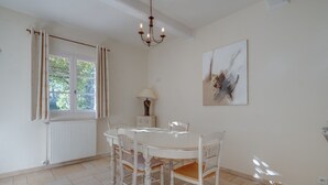 Un comedor con una mesa y sillas blancas, una araña, una ventana con cortinas, un radiador, una lámpara y un cuadro en la pared.