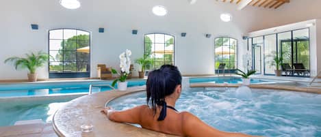 En kvinna som kopplar av i ett bubbelbad, med en pool och stora fönster i bakgrunden.