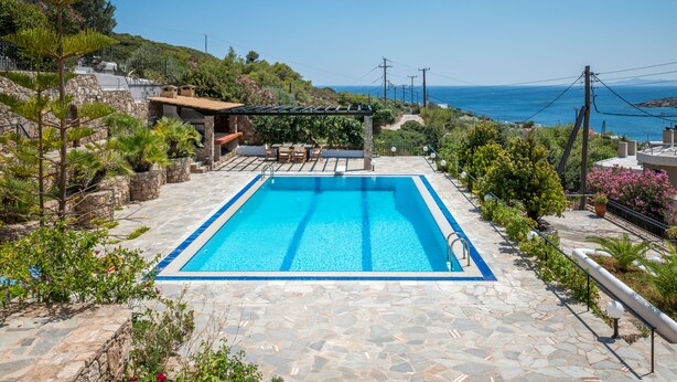 A rectangular swimming pool with clear blue water, surrounded by stone tiles and lush greenery. - Villa Barite (5 bedrooms) (Lavrio)