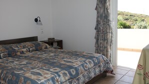 A bedroom with a patterned bedspread, a nightstand with books, a wall-mounted lamp, and a view of a hillside through a window. - Villa Barite (5 bedrooms) (Lavrio)