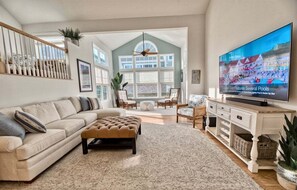 A spacious living room with a large TV, a sofa, an ottoman, and a view of the outside.