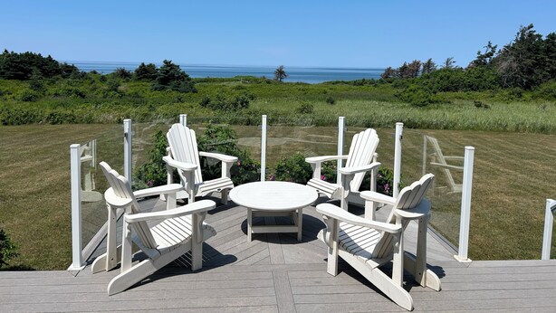 Une terrasse dotée de quatre fauteuils Adirondack blancs et d’une table ronde, offrant une vue sur une pelouse et l’océan.