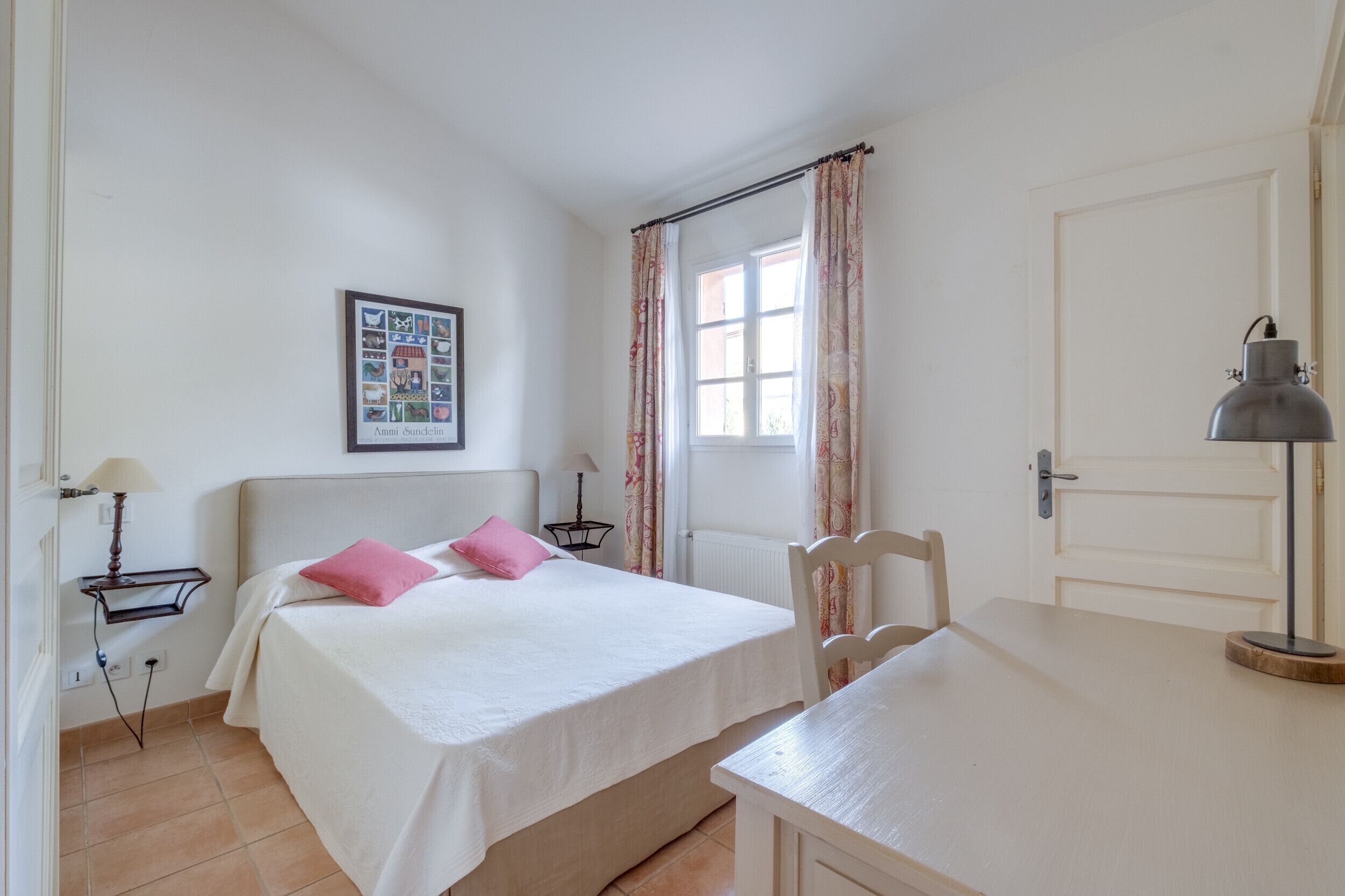 Ein Schlafzimmer mit Bett, Stuhl, Schreibtisch, Lampe, einem Bild an der Wand und einer Tür.