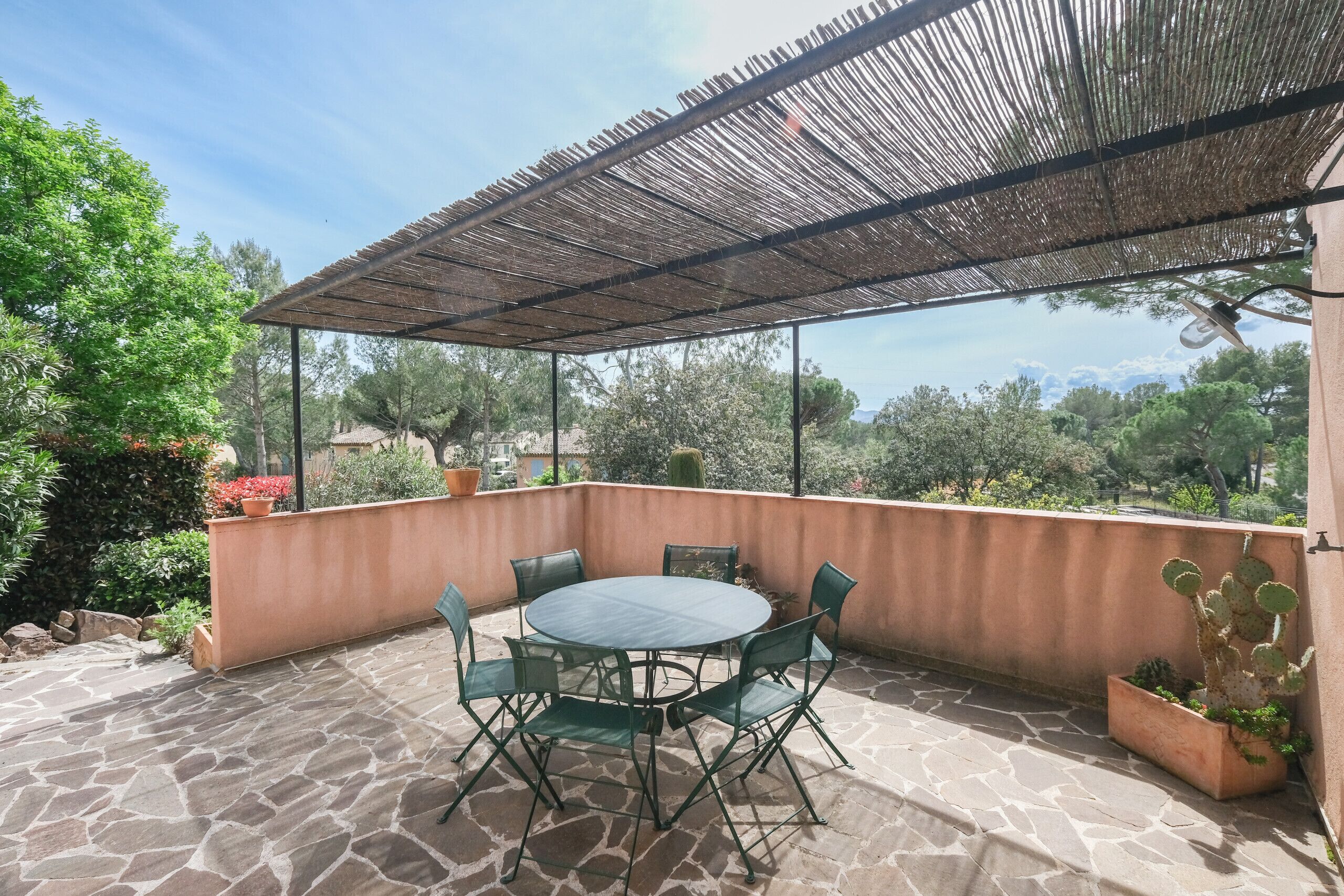 An outdoor patio with a round table and chairs, surrounded by potted plants and a pergola.