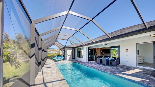 Une piscine intérieure moderne avec des parois en verre transparent, un espace patio doté de chaises longues et une cuisine visible à travers une porte ouverte.