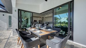Un área de comedor al aire libre con una mesa larga, sillas de mimbre, puertas correderas de vidrio y vistas del paisaje.