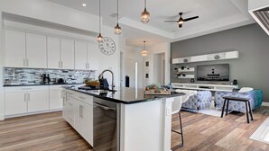 Una cocina moderna con una isla central, armarios blancos y un comedor con sofá gris y televisor.