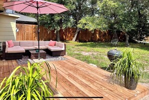 Ein Garten mit Holzterrasse, einem Ecksofa, einem roten Sonnenschirm und einem Grill.