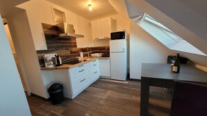 Una cocina moderna con armarios blancos, campana extractora de acero inoxidable, microondas integrado y refrigerador. Hay una mesa de comedor con una botella de vino y una cafetera. La cocina tiene un techo inclinado con tragaluces.