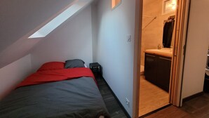 Un dormitorio con techo inclinado, cama con ropa de cama roja y gris, mesita de noche pequeña y puerta que da a un baño con espejo y lavabo.