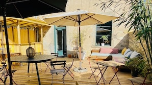 En udendørs patio med trægulv, et rundt bord med stole, en sofa og en stor parasol, der giver skygge.