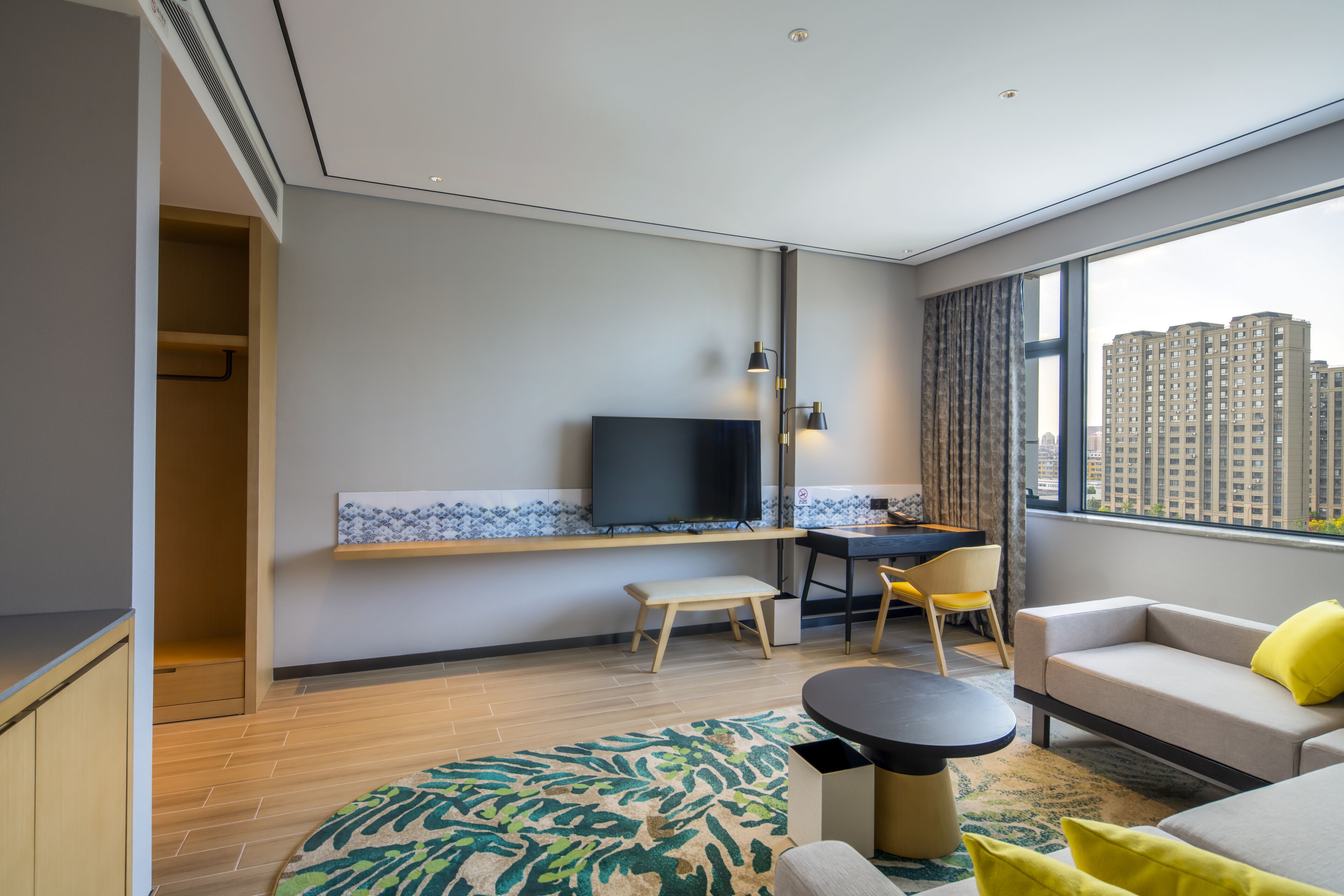 A modern hotel room with a large window, a flat-screen TV, a desk, a sofa, and a small table.