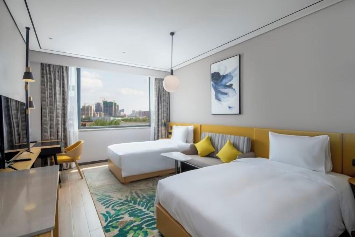 A hotel room with two beds, a desk, a chair, and a large window offering a city view.