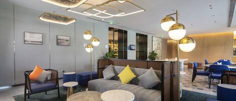 A modern hotel lobby with a sofa, chairs, and tables, featuring geometric ceiling designs and ambient lighting.