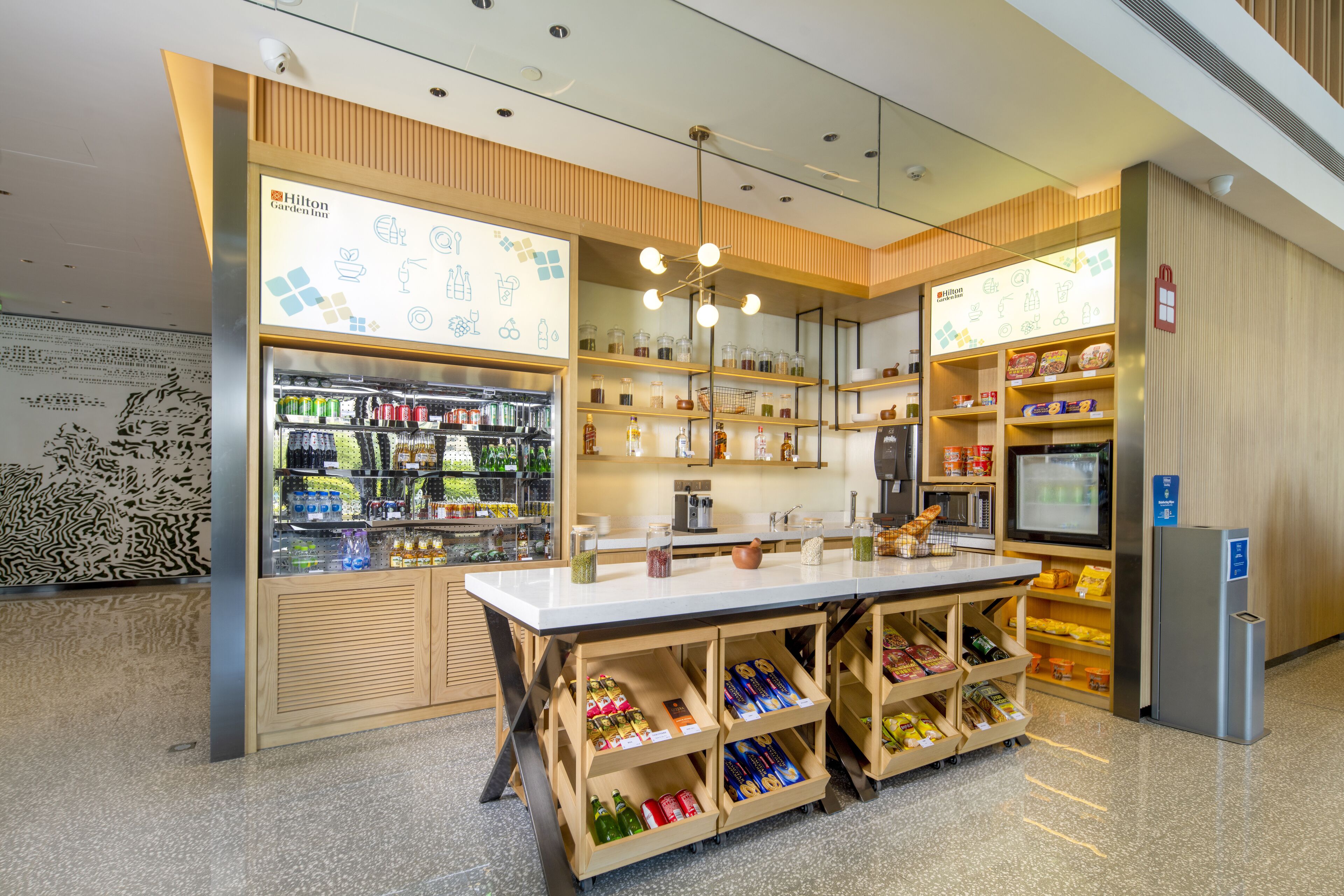 A well-lit hotel lobby with a self-service beverage station, a television, and a refrigerator.