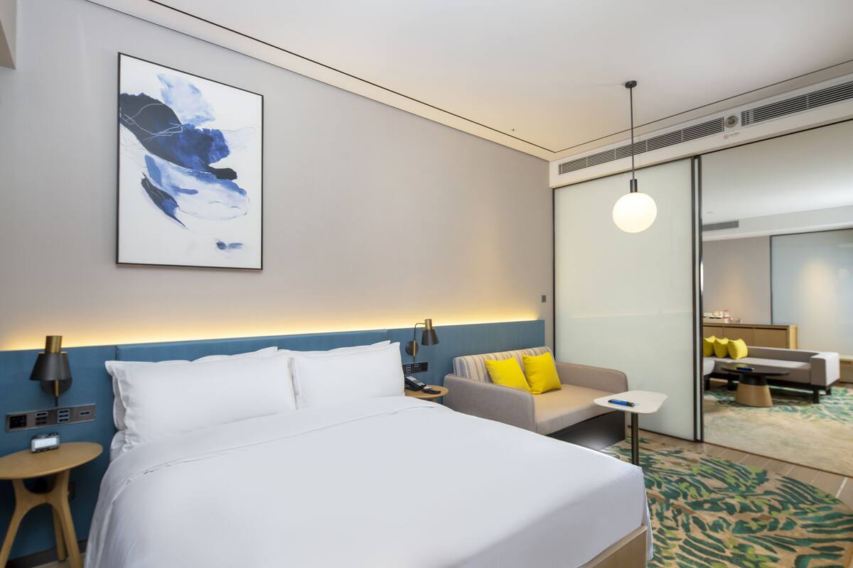 A modern hotel room with a large bed, a sofa, a small table, and a wall-mounted artwork.