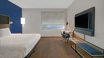 A hotel room with a bed, a desk, a chair, a bench, a TV, and a window with blinds.