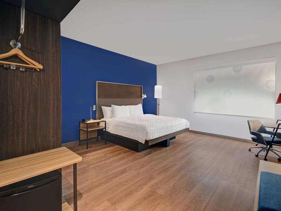 A modern hotel room with a bed, bedside table, chair, and a desk with a chair.
