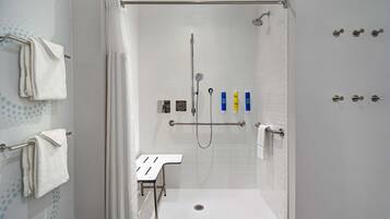 A walk-in shower with white tiled walls, a showerhead, a handheld showerhead, and a grab bar. There are towels on a rack and a small table with a towel.