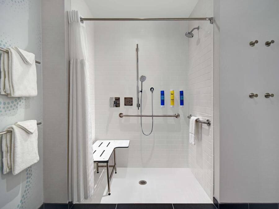 A walk-in shower with white tiled walls, a showerhead, a handheld showerhead, and a grab bar. There are towels on a rack and a small table with a towel.