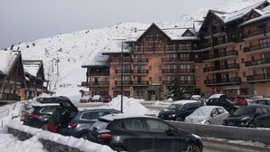A snow-covered parking area with several cars and a stone wall. In the background, there are multi-story buildings with balconies and a ski lift visible on the mountain. - 5pers. Running boards Modern Mountain View (Valmeinier)