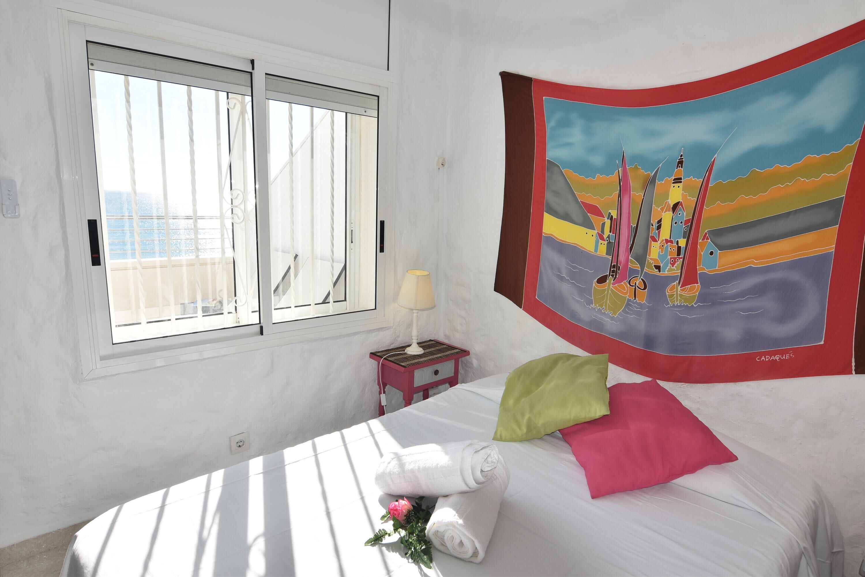 A hotel room with a bed, a window, a nightstand, and a colorful tapestry on the wall.