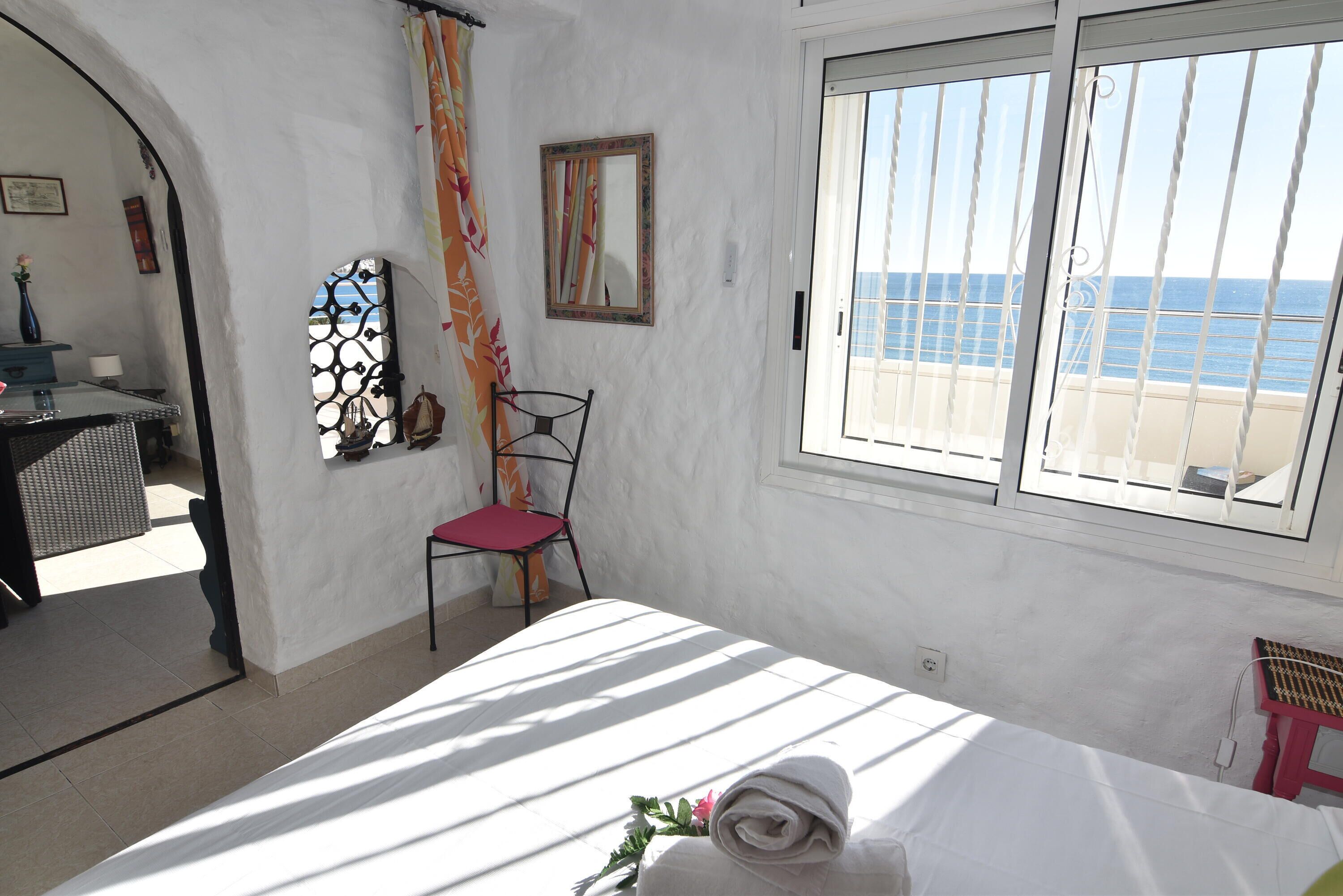A bedroom with a bed, a chair, a window with a view of the sea, and a door leading to another room.