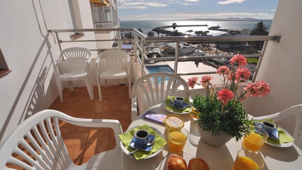Een balkon met witte plastic stoelen, een tafel gedekt met ontbijtartikelen, een bloevas en uitzicht op zee en gebouwen.
