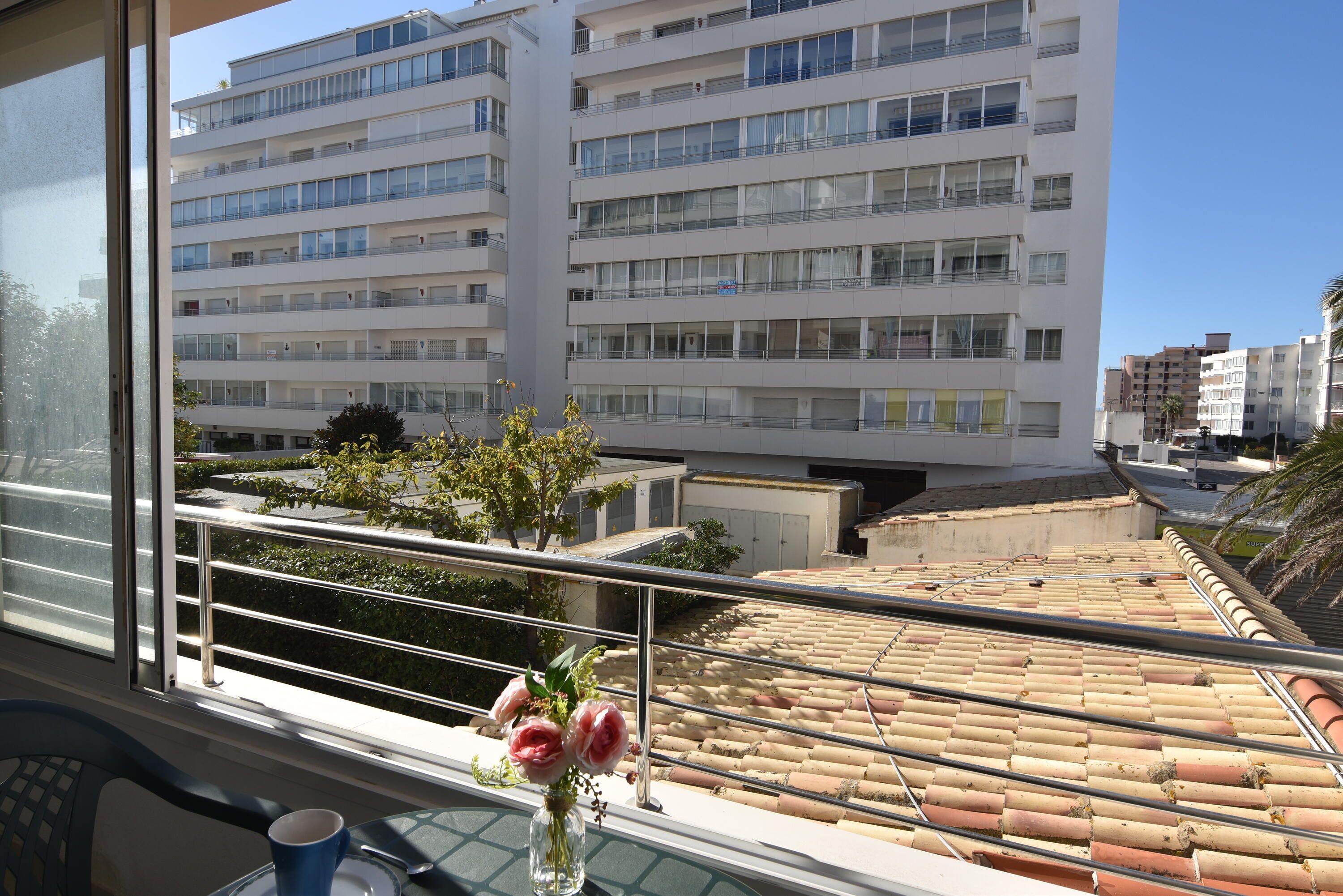 Ein Balkon mit Blick auf ein mehrstöckiges Gebäude, eine Vase mit Blumen und ein Tisch mit einer Tasse.