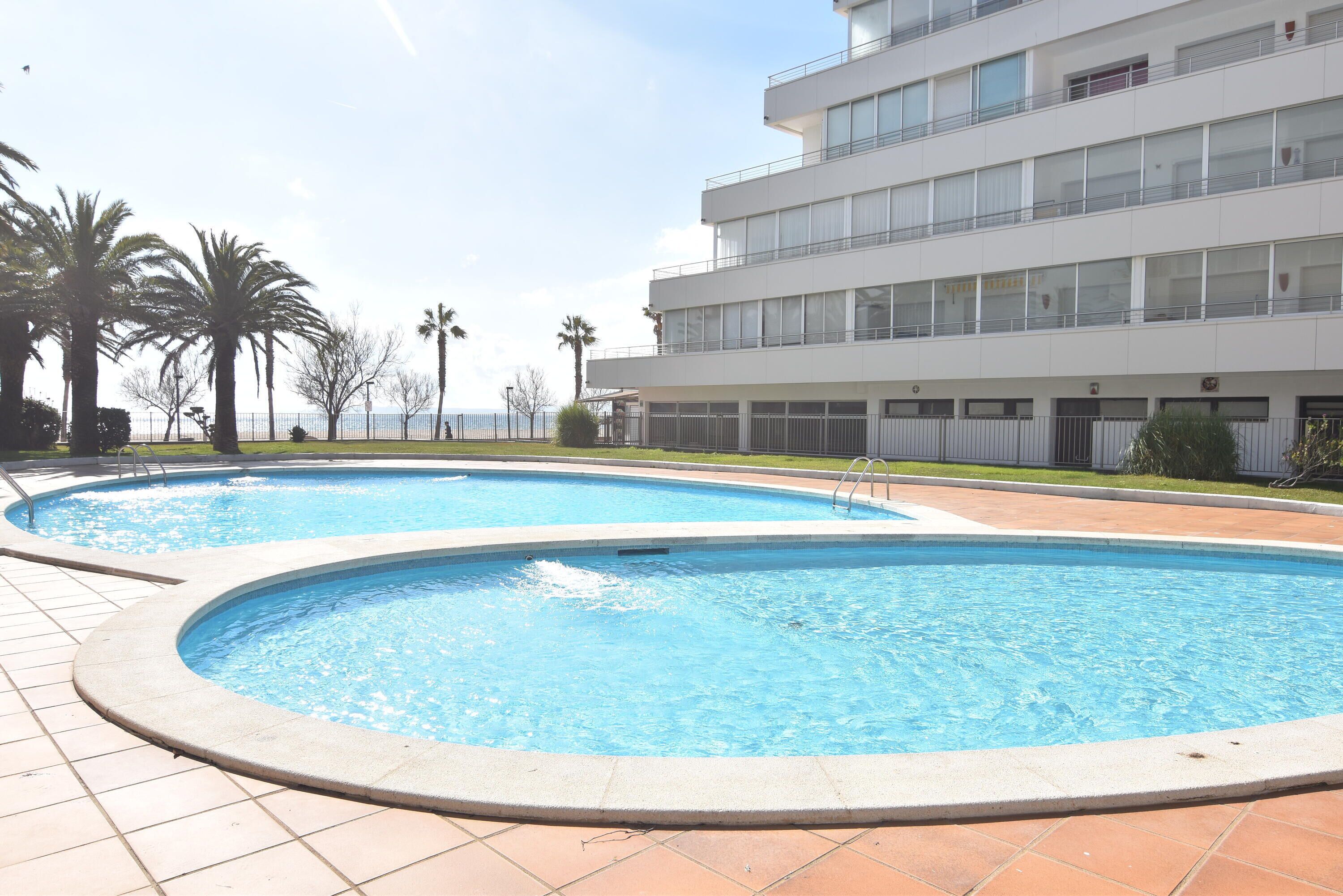 A swimming pool area with two pools, surrounded by palm trees and a modern building with multiple balconies.