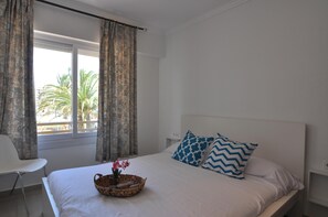 A bedroom with a bed, white curtains, and a window with a view of palm trees. - Charming apartment for rent in Roses, Costa Brava! 0 meters from the beach. Enjoy the community pool! (Roses)