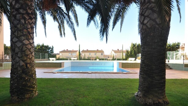 A swimming pool with a sign reading "EDMEDITERRANI 1_2" surrounded by palm trees and a grassy area. - Apartment in Costa Brava, 2 bedrooms for 6 people, mountain views, community pool, close to beach, supermarket and restaurants. (Roses)