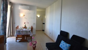 A dining area with a table set for four, a black sofa with a patterned pillow, and a wall-mounted air conditioner. - Apartment in Costa Brava, 2 bedrooms for 6 people, mountain views, community pool, close to beach, supermarket and restaurants. (Roses)