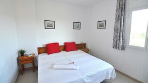 A neatly made bed with white linens and red pillows, a small wooden nightstand with a plant, and a window with patterned curtains. - Apartment in Costa Brava, 2 bedrooms for 6 people, mountain views, community pool, close to beach, supermarket and restaurants. (Roses)