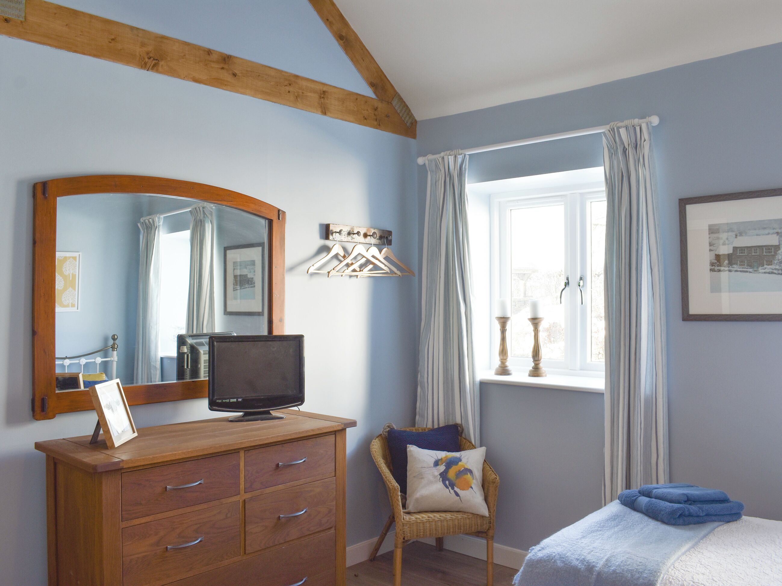 A bedroom with a wooden dresser, a television, a chair with a cushion, a window with curtains, and a bed with a blue blanket.