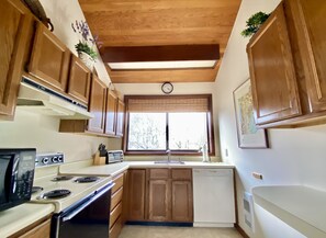 A compact kitchen with wooden cabinets, a stove, a dishwasher, and a window with a view of trees.