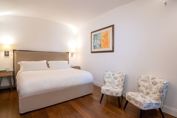 A bedroom with a large bed, two chairs, a nightstand, and a wall-mounted lamp.