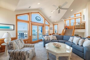A spacious living room with a large sectional sofa, a glass coffee table, and a view of the outside. - Acqua - KDH MP 6.5, Ocean View, Steps to Beach, Elevator, Pool, Hot Tub, Rec Room (Kill Devil Hills)