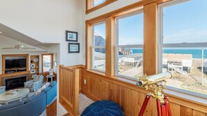 Una sala de estar con televisor, sofá y un telescopio junto a un ventanal con vista a la playa.